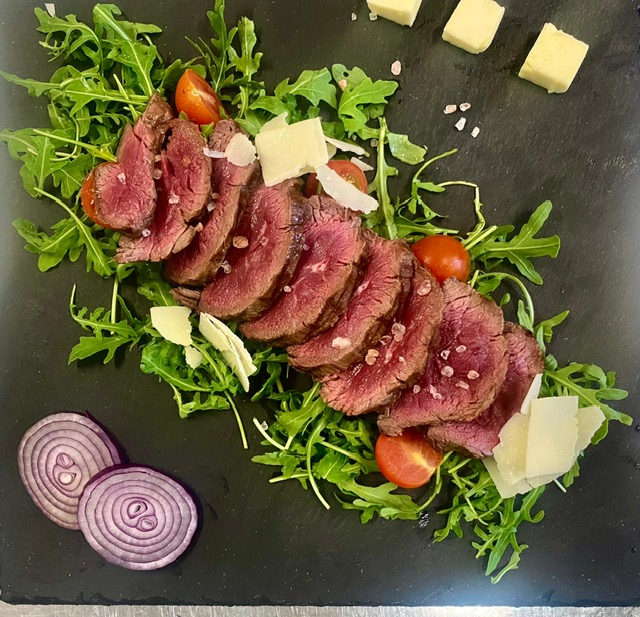 Dunkle Fleischscheiben auf Rucola mit Tomaten und Käsestücken, rote Zwiebeln daneben